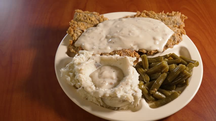Close-up view of chicken fried steak smothered in creamy country gravy, served with mashed potatoes and green beans, with warm lighting and rustic table setting. Slider, 4K