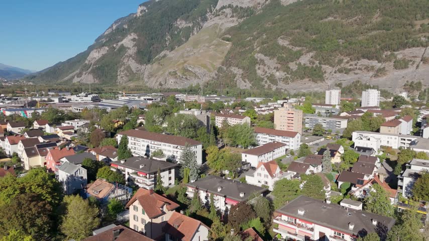 Mixed residential buildings and tall apartment towers spread across valley floor beneath steep forested mountains and rocky cliffs under clear sky in Chur, Switzerland. Aerial drone orbit wide shot