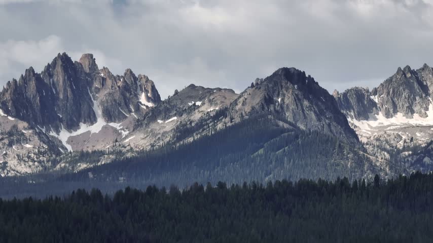 Stanley Idaho Sawtooth Mountains feature dramatic rocky peaks with snow patches rising above a dense pine forest under a cloudy sky in this scenic landscape view