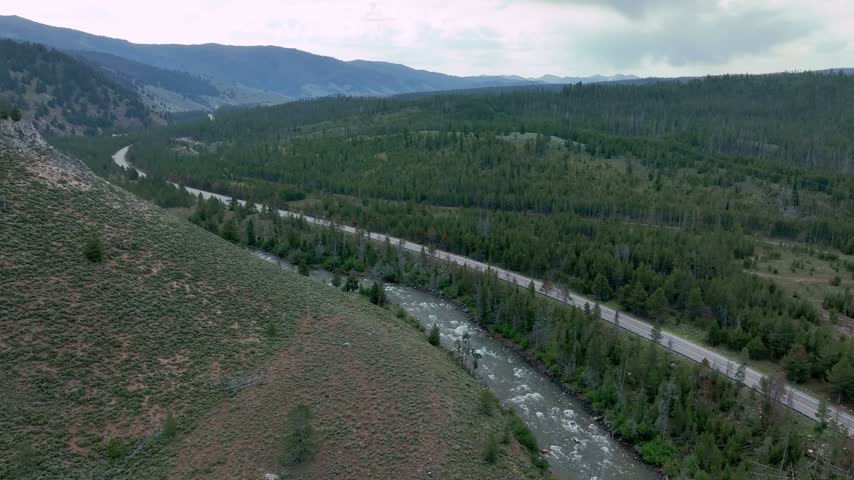 Aerial view shows vehicles driving on a mountain highway beside a winding river through dense pine forests in Stanley Idaho