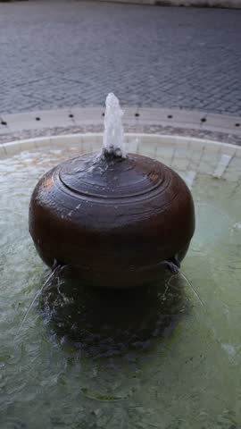 Vertical screen bronze spherical fountain sprays water upward and streams flow from side spouts into a circular stone basin creating gentle ripples in a quiet public square
