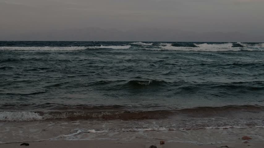 Low-angle view of small waves breaking onto sandy shore under overcast sky, frothy whitewater washing across wet sand with distant hazy mountain silhouette