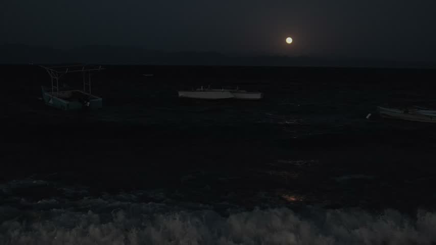 Full moon over rough ocean waves at night, boats rocking on the water and moonlight reflections creating a dramatic cinematic seascape