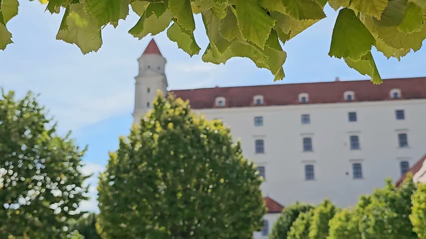 Bratislava Castle in Slovakia viewed from the surrounding gardens, showing the historic white castle building and tower framed by trees under a blue sky.