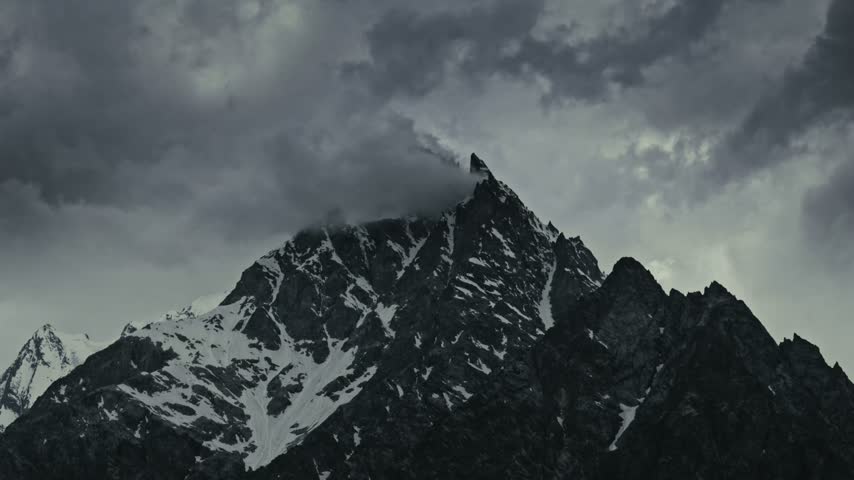Timelapse of a majestic snow covered mountain peak beneath dramatic storm clouds Flowing through the sky, with rugged rocky slopes, snow streaks and glaciers contrasting against the dark sky..