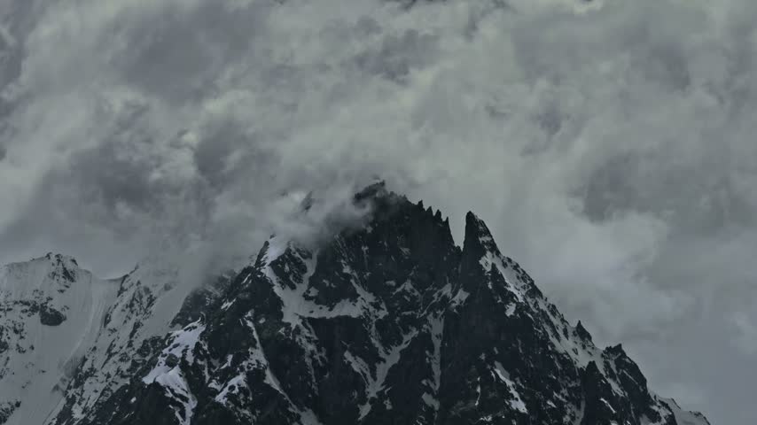 Timelapse of a majestic snow covered mountain peak beneath dramatic storm clouds Flowing through the sky, with rugged rocky slopes, snow streaks and glaciers contrasting against the dark sky..