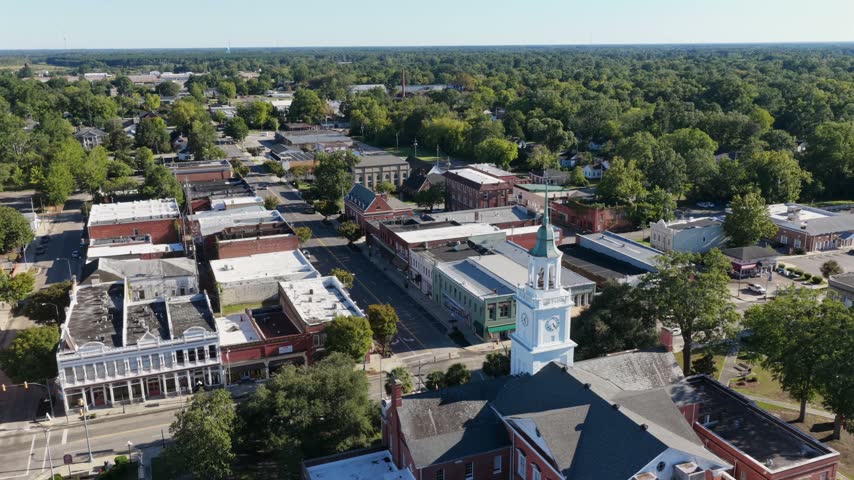 Brick courthouse with a gray roof and white clock tower sits among oak trees beside storefronts and a green lawn in Bennettsville, South Carolina, USA. Aerial view