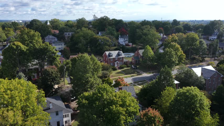 Red brick colonial house with dormer windows, a white porch and a gray slate roof stands behind green and orange trees as the view closes in, Lynchburg, Virginia, USA. Aerial approaching shot