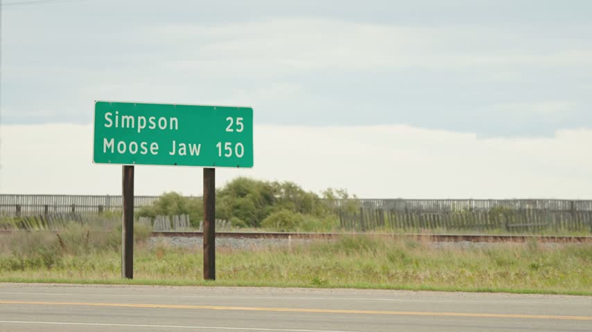 Static shot of a green and white provincial highway sign near Watrous, Saskatchewan, showing distances to Simpson and Moose Jaw with grass, train tracks and an overcast sky behind