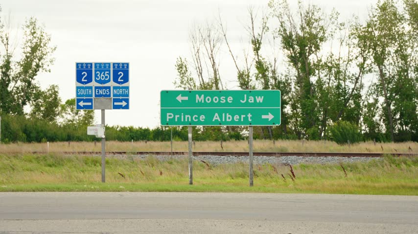 Static shot of a green and white provincial highway sign outside Watrous, Saskatchewan, with arrows to Moose Jaw and Prince Albert, bushes and grass behind and an intersection in front