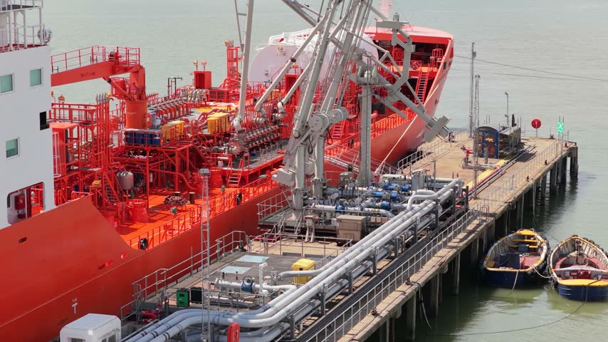 A large orange tanker vessel is docked at an industrial port terminal with several pipes and cranes for loading operations while small boats float in the water nearby.