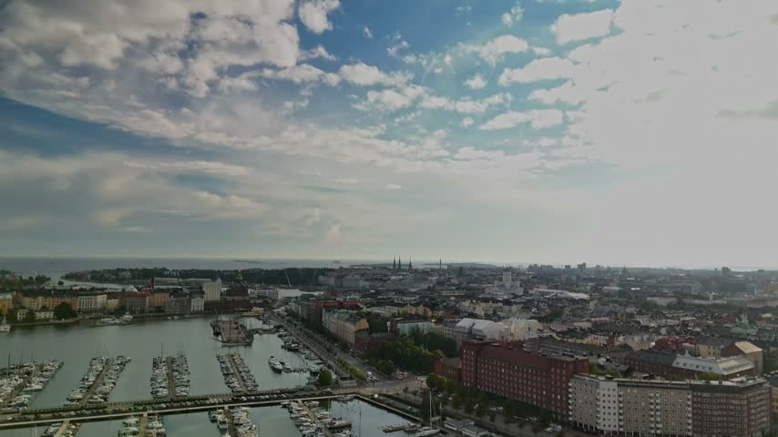 Drone view across a Helsinki marina in Finland where many sailing yachts and motor boats are berthed along extended piers with old city buildings and cloudy sky beyond