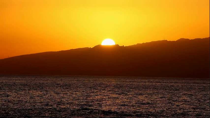 Sunset over the horizon, with mountains rising above the calm sea against an orange sky.