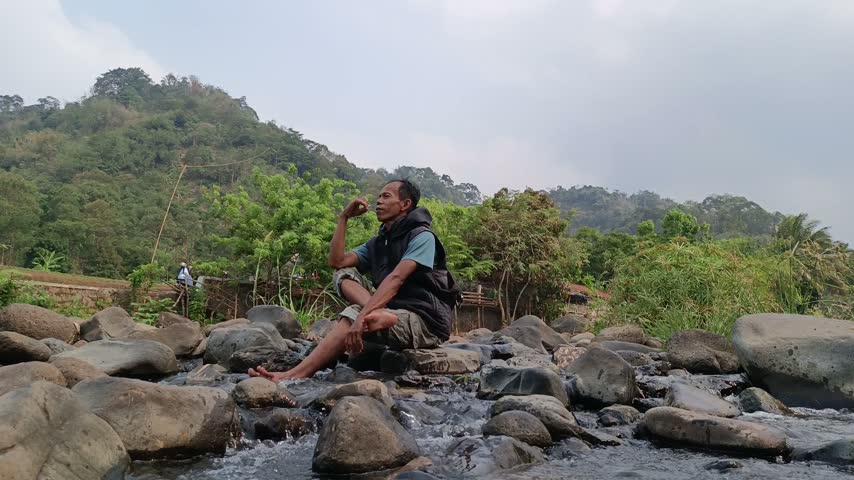 Relaxed Man Sitting On Boulders In A Flowing Stream