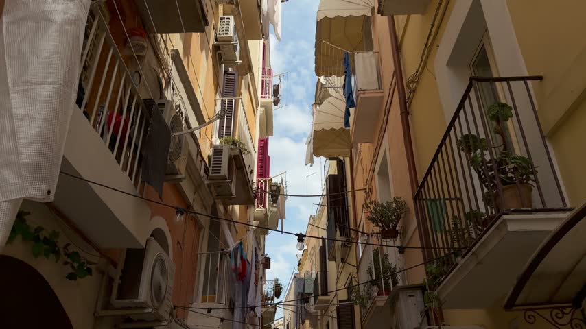 Slow forward view up a narrow street in Bari old town, Puglia, Italy, with hanging laundry, iron balconies and ochre facades, tilting up toward the sky