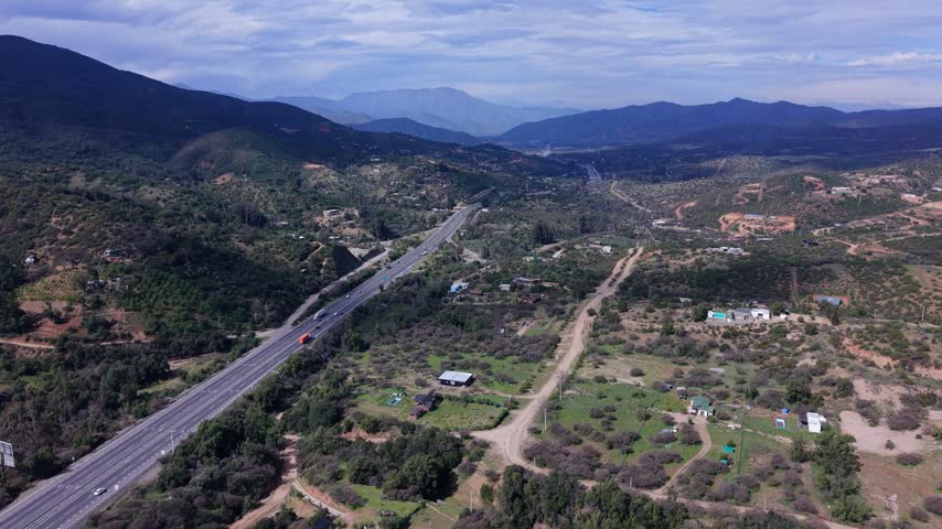 Aerial view of a highway winding through a rural valley with scattered farmhouses, a swimming pool and dirt roads under a cloudy sky