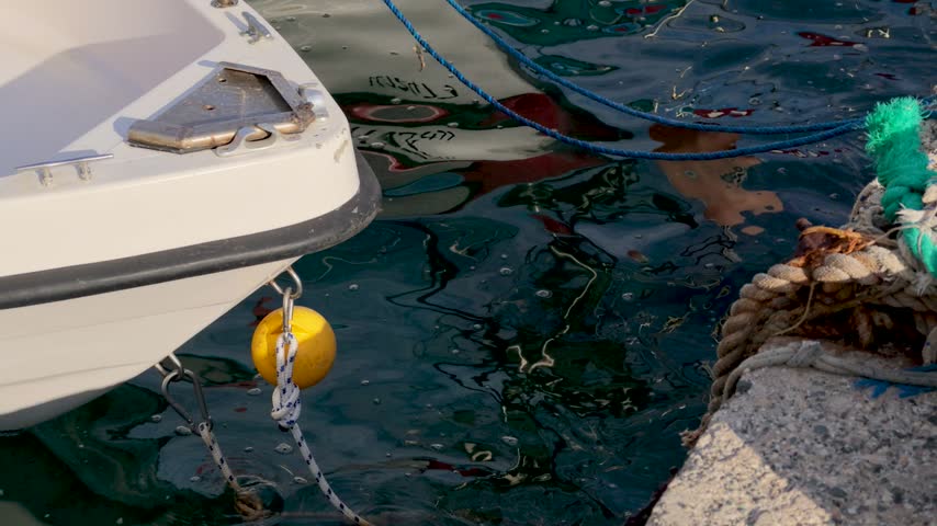 A boat is secured at the harbor with a yellow buoy floating nearby. The mooring line connects the boat to the dock. Water reflects the surroundings clearly.