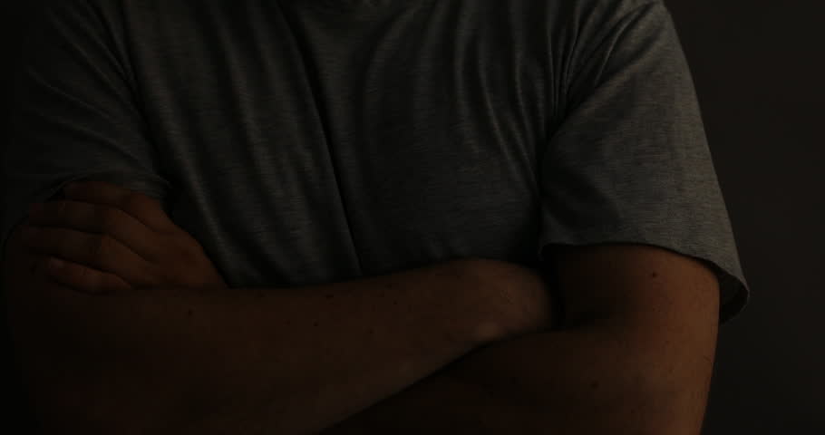Unrecognizable person in casual attire standing with crossed hands in shadow.