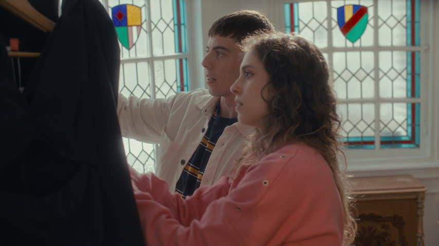 Young man and woman smiling and talking as inspecting black dress on hanger, standing by stained glass window inside vintage secondhand shop