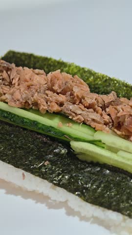 Hands spreading grilled salmon filling over cucumber and cream cheese on a nori sheet, sushi preparation