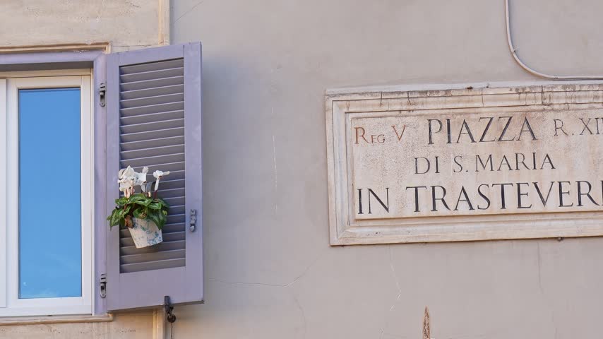 Close up view of the ancient marble street sign of Piazza di Santa Maria in Trastevere, next to a window with a traditional purple wooden shutter and a small plant, in Rome, Italy