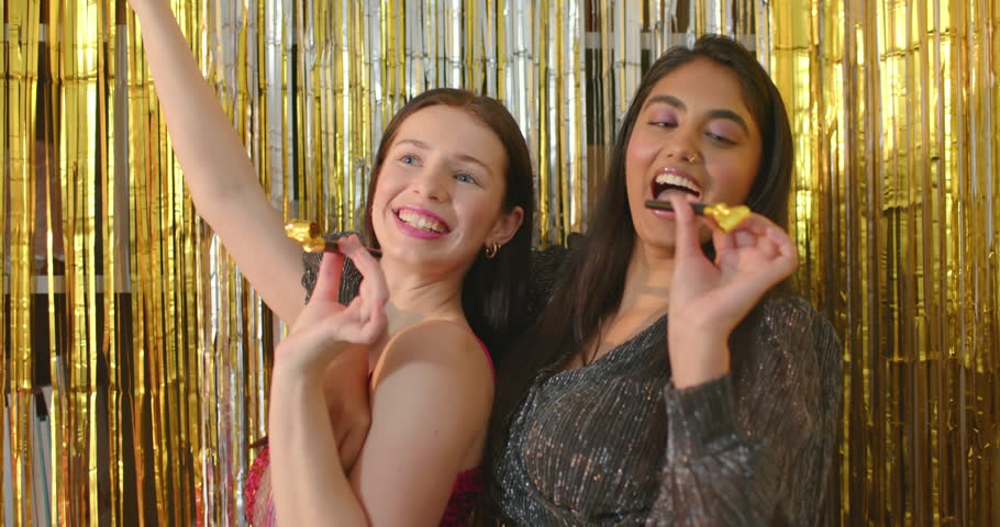 Two adult women in sequin dresses celebrating hugging blowing gold horns before gold curtain. Friends, celebration, glam, fringe, blowers, studio, gowns