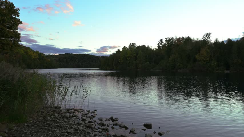Soft evening colors brighten the sky above calm lake water and a dark tree lined shore at Green Lakes State Park, New York.