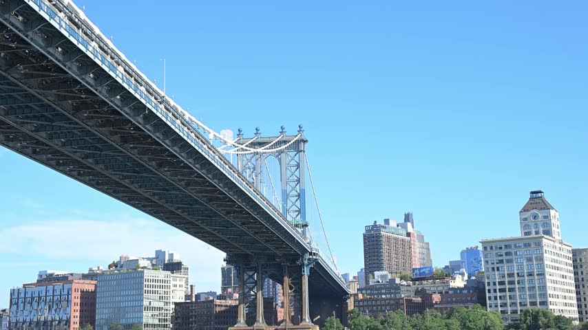 An upward waterfront angle looks beneath the Manhattan Bridge roadway toward its tower and nearby city buildings.