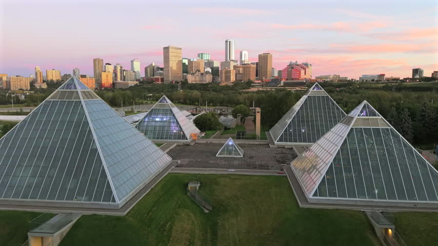 Forward drone flight passes between the Muttart Conservatory glass pyramids and over the transit corridor toward downtown Edmonton in soft morning light.