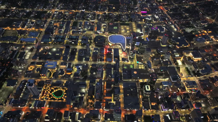 Retreating high aerial reveals downtown Edmonton at night, with a brightly illuminated arena, dense city blocks, glowing streets and moving traffic below.