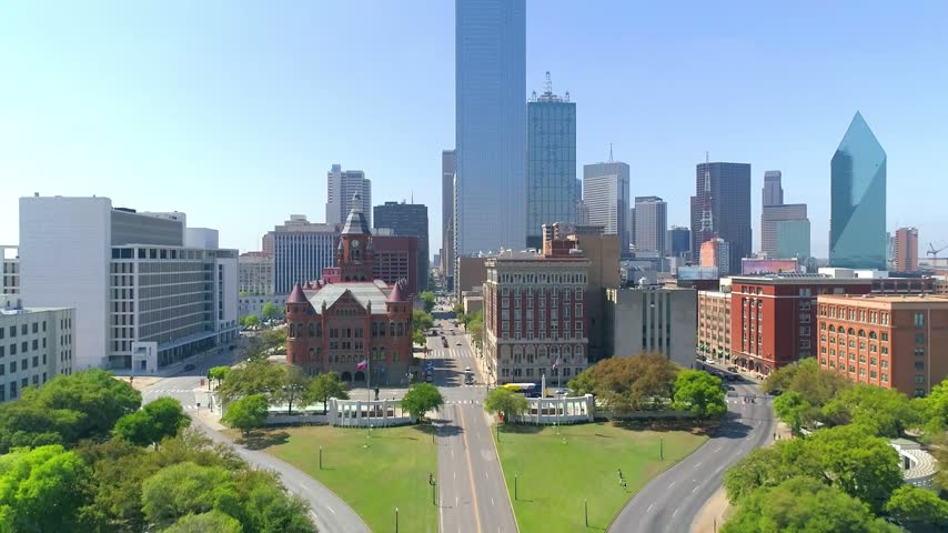 High-angle scenic aerial drone view looking east over the historic Dealey Plaza park and Elm Street corridor, showcasing the red stone Dallas County Courthouse (Old Red Museum), Dealey Plaza Grassy Knoll, and towering skyscrapers of the Downtown Dallas central business district.
