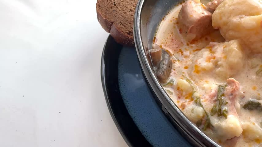 Close view of creamy chicken and vegetable soup with dumplings, mushrooms, leafy greens, and bread beside a dark bowl as the camera gradually reveals the hearty meal