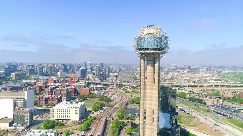 Wide high-angle aerial drone perspective highlighting the geometric sphere of Reunion Tower, showcasing Dealey Plaza, the historic red stone Dallas County Courthouse, winding railroad tracks, and sprawling corporate buildings of Dallas, Texas, under a clear sky.