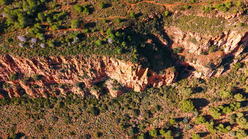 Aerial view of mountain path along red cliff edge. Top down shot showing a walking trail through green forest next to a steep rocky drop.