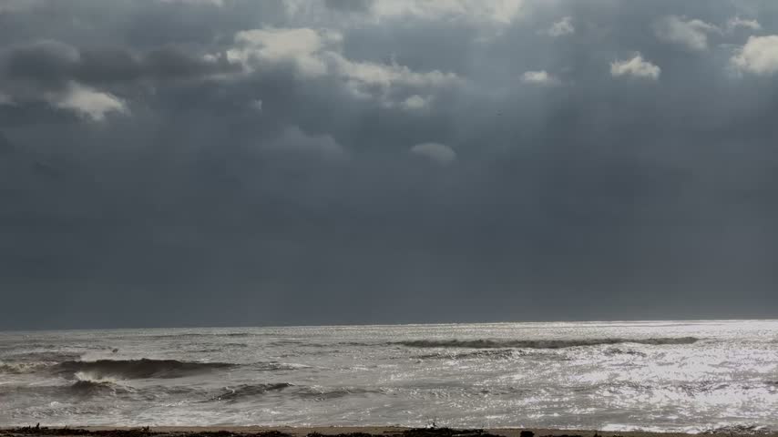 Slow motion rough sea with breaking waves under dark storm clouds and sun rays, silver sunlight glittering on the water surface, dramatic stormy seascape with copy space