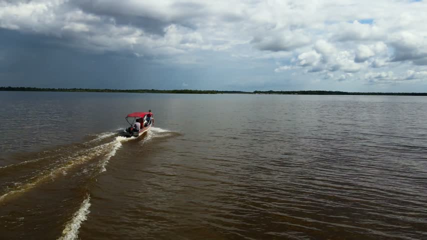Aerial view of a small boat with a red canopy navigating the dark waters of the Amazon River near Manaus, Amazonas, Brazil.