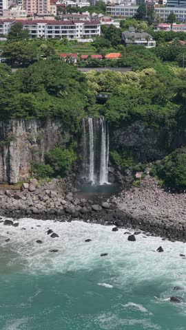 Jeongbang Waterfall Cascading Down A Rocky Cliff Into The Sea In Seogwipo, Jeju Island, South Korea. - aerial vertical shot
