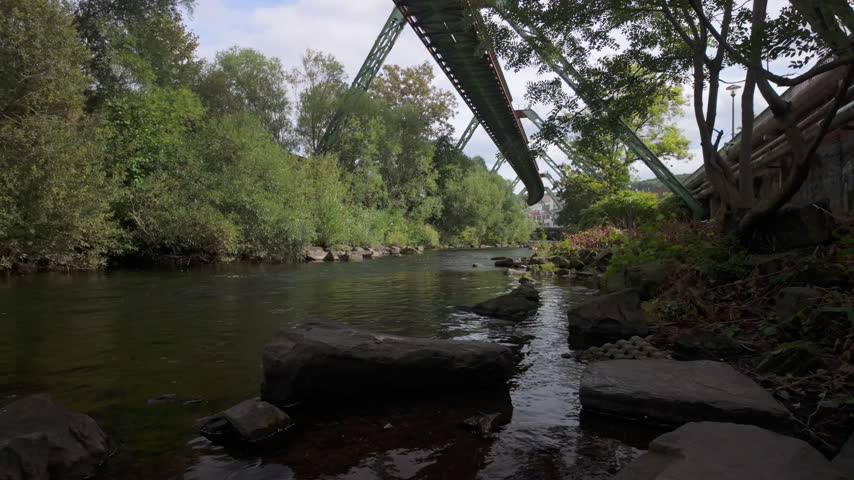 Wuppertal Schwebebahn train departs above the Wupper River in Germany, surrounded by green trees and rocks from a scenic low riverside viewpoint.