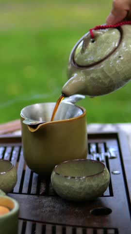 Tea flows from handled ceramic pot into cup on tea table outdoors. Tea service features matching small bowls and garden greenery, suggesting quiet moment of traditional hospitality.