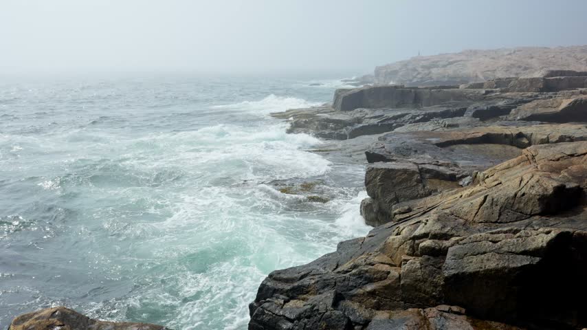 Crashing ocean waves along the rugged pink granite rock ledges enveloped in thick fog at Schoodic Point in Acadia National Park, Maine.