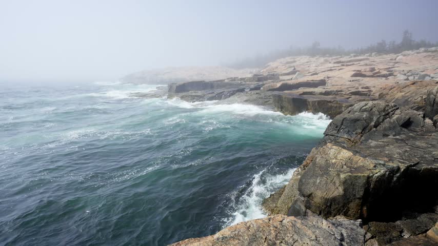 Crashing ocean waves along the rugged pink granite rock ledges enveloped in thick fog at Schoodic Point in Acadia National Park, Maine.