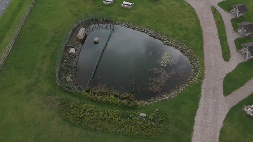 Aerial view of a small pond with a wooden enclosure surrounded by lush green grass and a gravel path in Saare County, Estonia.