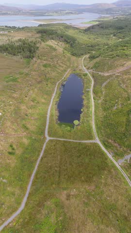 Aerial view of a small pond surrounded by green hills and walking trails in the rural landscape of Bonny Glen Woods Portnoo, County Donegal, Ireland.