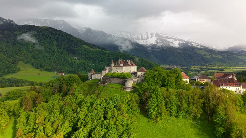 Medieval Gruyeres Castle on a green hill with the Swiss Alps in the background under a cloudy sky.