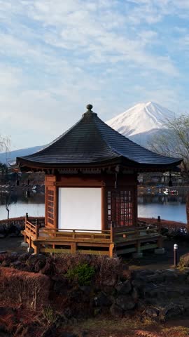 Aerial panning view of iconic hexagonal wooden gazebo by tranquil water surface near snow capped Mount Fuji landmark.