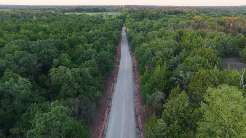 Empty dirt country road cutting through a dense green tree canopy in an aerial view over rural Oklahoma.