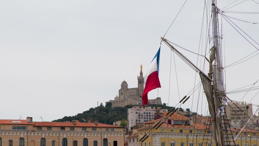 French tricolor flag flutters from a ship rigging overlooking Marseille and Notre-Dame de la Garde.