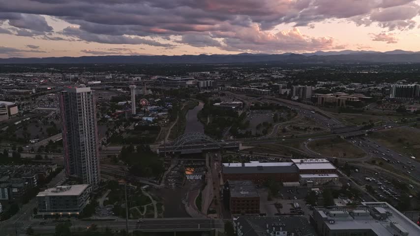 Aerial view of Denver skyline with the South Platte River, urban infrastructure, and distant mountains under a dramatic sunset sky in Denver, Colorado, United States.