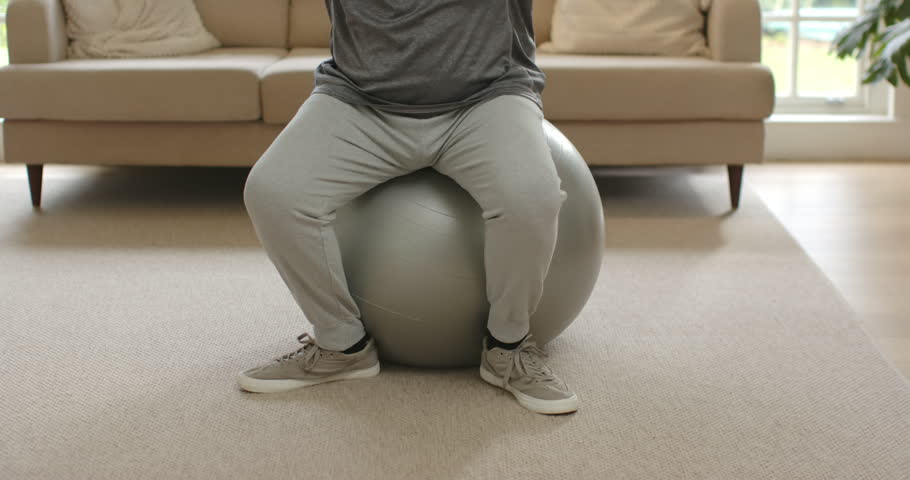 African American man in activewear practicing balance on silver ball leaning, sweeping arms at home. Stabilityball, livingroom, sofa, daylight, window, plant, fitness