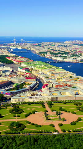 Vertical video. Panoramic aerial view of the city center of St. Petersburg, Russia. The footage opens over the Field of Mars, a nearly nine-hectare open park that has served as a military parade ground, festival space, and revolutionary memorial since the early 18th century. The camera sweeps across the geometric lawns and pathways of the Field of Mars, bordered by the Moyka River to the south and the Swan Canal to the east. Visible nearby are the distinctive green roofs of the Mikhailovsky Castle and the golden spire of the Admiralty building rising above the dense urban fabric. The wide Neva River cuts through the historic center, with the Trinity Bridge and distant cable-stayed bridges visible against the horizon. The cityscape is composed of baroque and neoclassical palaces, government buildings, and orthodox churches with onion domes, all bathed in bright daylight under a cloudless blue sky. The scene captures the harmonious ensemble of St. Petersburg